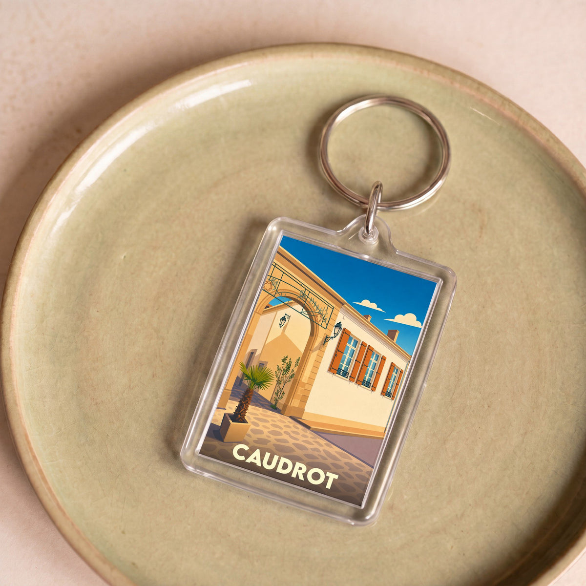 Porte-clés souvenir de la ville de Caudrot avec une illustration stylisée d'une rue ensoleillée.