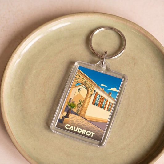 Porte-clés souvenir de la ville de Caudrot avec une illustration stylisée d'une rue ensoleillée.