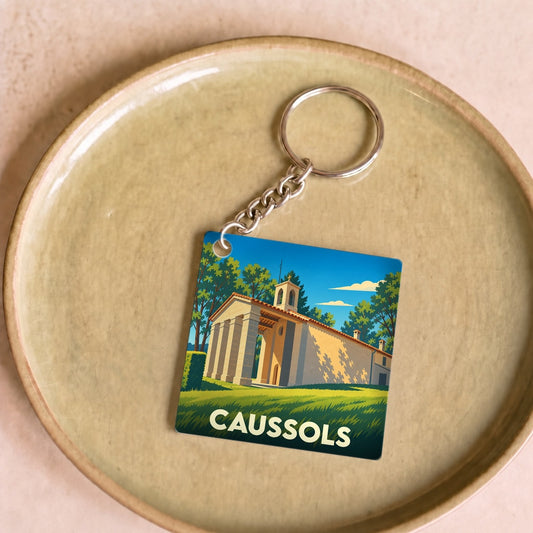 Porte-clés souvenir de la ville de Caussols avec une illustration stylisée d'un bâtiment provençal.