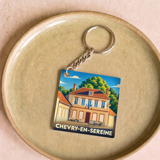 Porte-clés souvenir de la ville de Chevry-en-Sereine illustrant une bâtisse traditionnelle sous un ciel bleu.