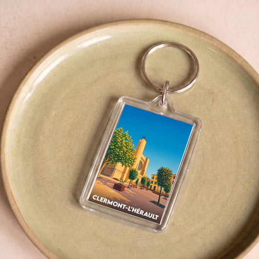 Porte-clés souvenir de la ville de Clermont-l'Hérault avec une illustration stylisée de son église et de ses arbres.