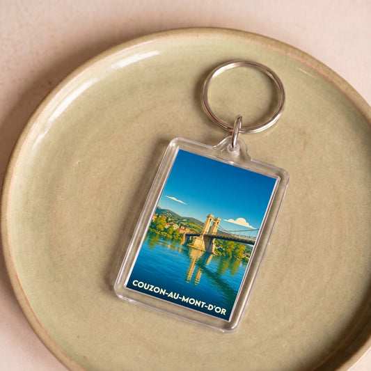 Porte-clés souvenir de la ville de Couzon-au-Mont-d'Or avec une illustration stylisée d'un pont suspendu sur une rivière.