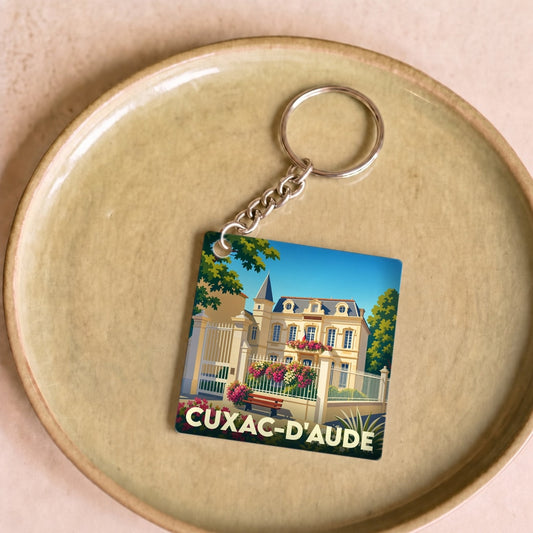 Porte-clés souvenir de la ville de Cuxac-d'Aude avec une illustration stylisée d'un bâtiment historique fleuri.