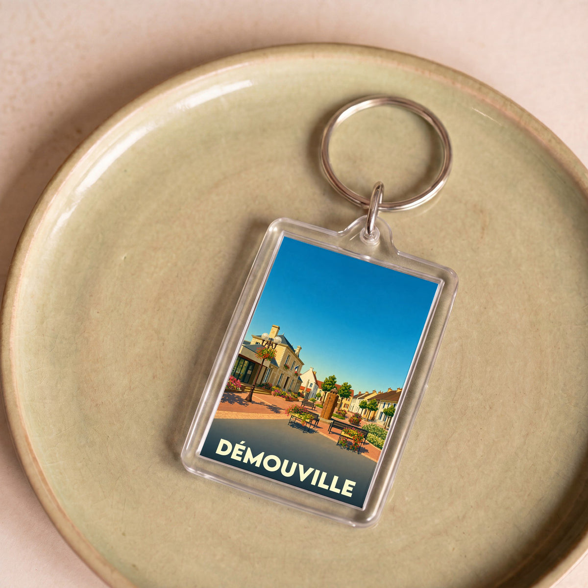 Porte-clés souvenir de la ville de Démouville avec une illustration stylisée de sa place principale.