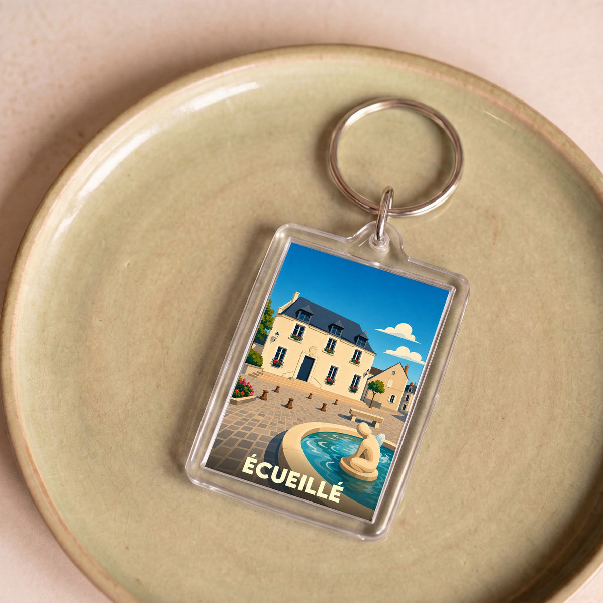 Porte-clés souvenir de la ville d’Écueillé avec une illustration stylisée d'une place et sa fontaine.