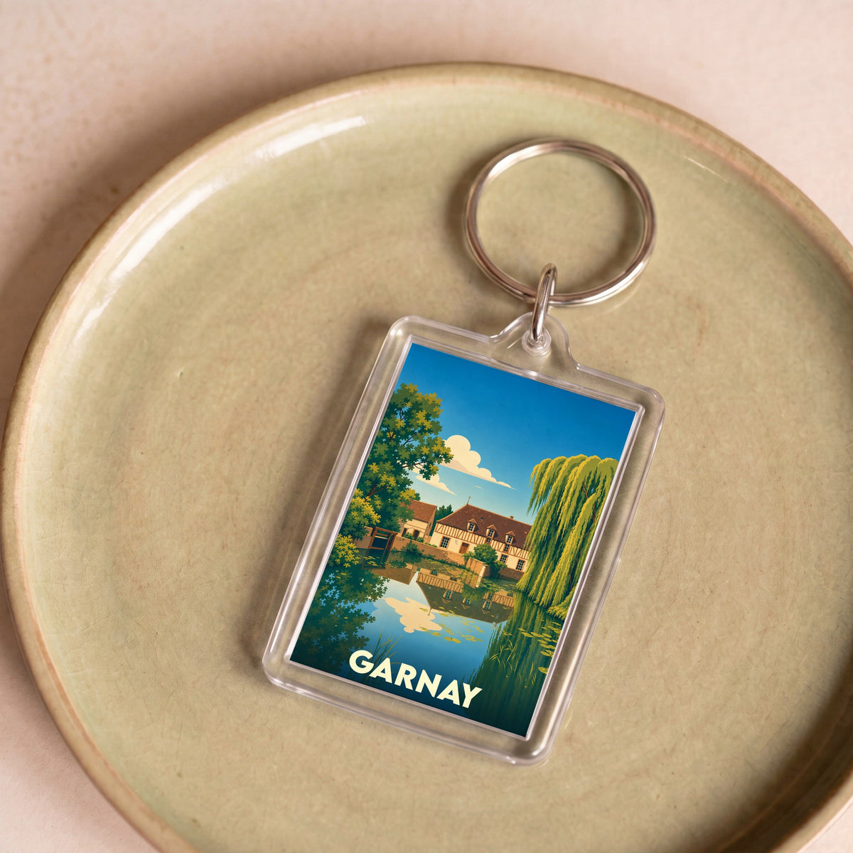 Porte-clés souvenir de la ville de Garnay avec une illustration stylisée d'un paysage bucolique au bord de l'eau.