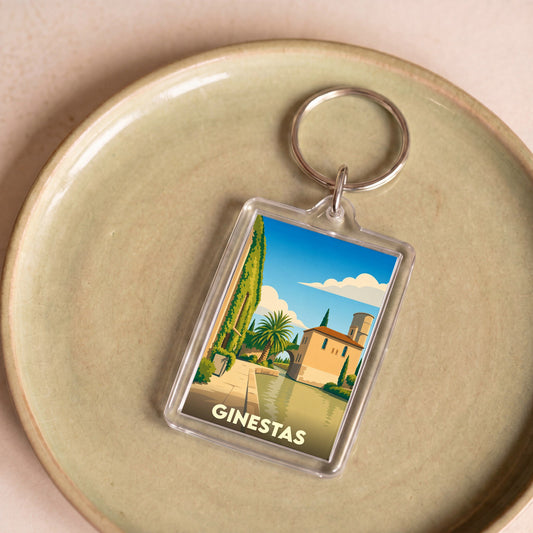 Porte-clés souvenir de la ville de Ginestas avec une illustration stylisée d'un paysage ensoleillé au bord de l'eau.