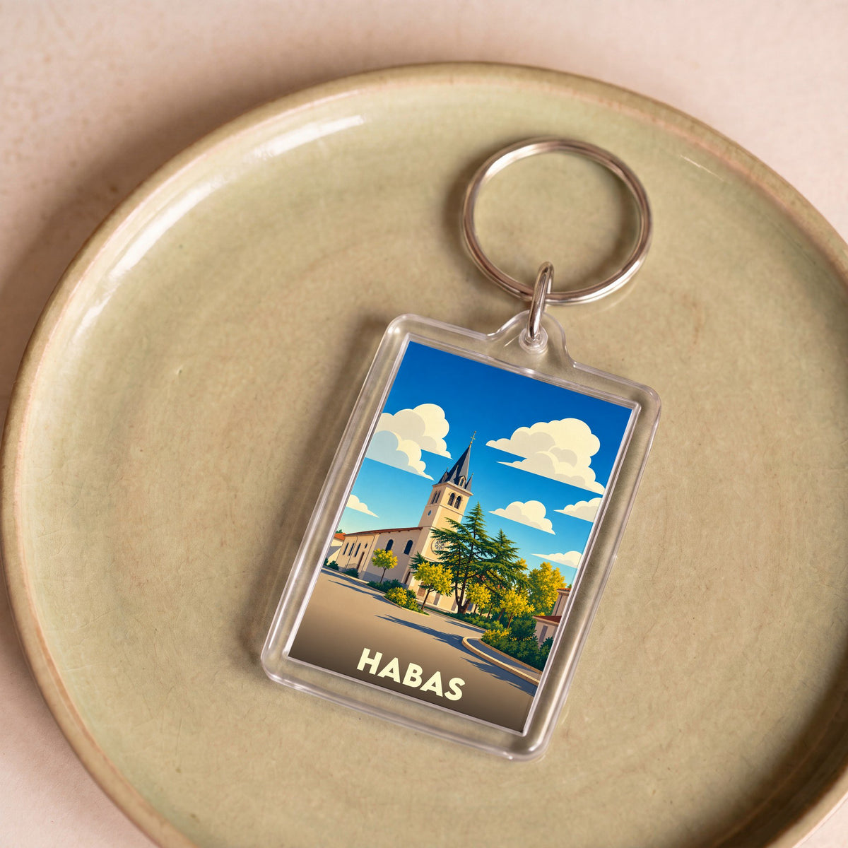Porte-clés souvenir de la ville de Habas avec une illustration stylisée de son église sous un ciel nuageux.