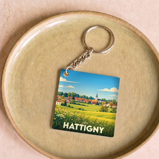 Porte-clés souvenir de la ville de Hattigny avec une illustration stylisée de son paysage champêtre.