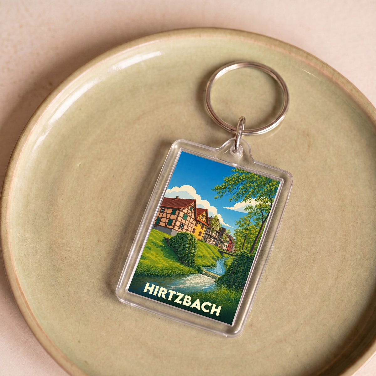 Porte-clés souvenir de la ville de Hirtzbach avec une illustration stylisée de maisons à colombages au bord de l'eau.
