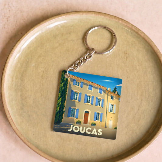 Porte-clés souvenir de la ville de Joucas illustrant une maison provençale aux volets bleus.