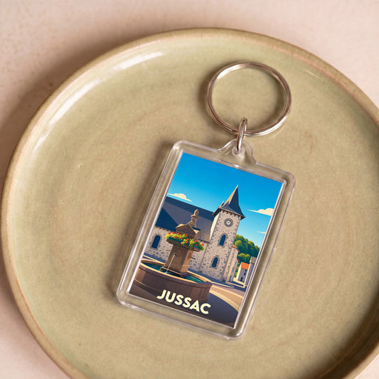 Porte-clés souvenir de la ville de Jussac avec une illustration stylisée d'une fontaine et de son église.