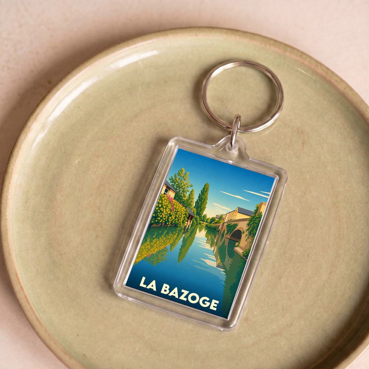 Porte-clés souvenir de la ville de La Bazoge avec une illustration stylisée d'un paysage fluvial.