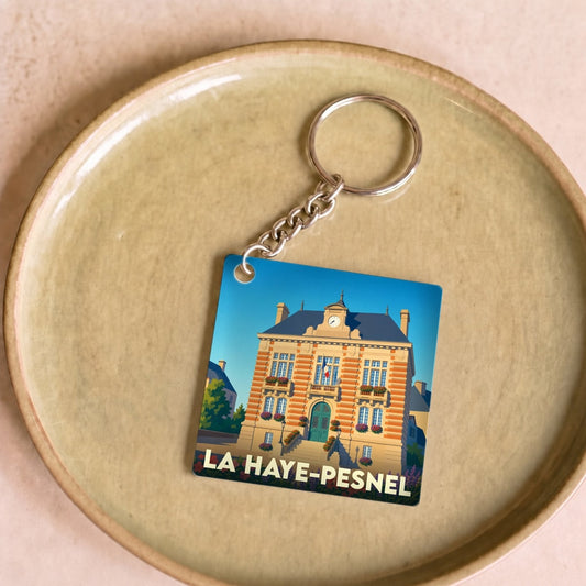 Porte-clés souvenir de la ville de La Haye-Pesnel avec une illustration stylisée de sa mairie fleurie.