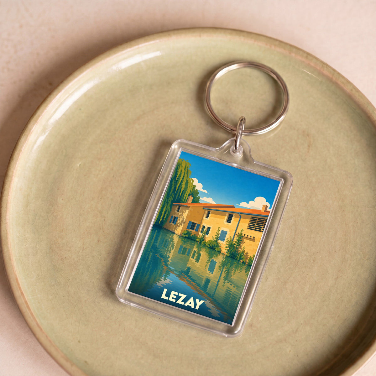 Porte-clés souvenir de la ville de Lezay avec une illustration colorée et stylisée de maisons au bord de l'eau.