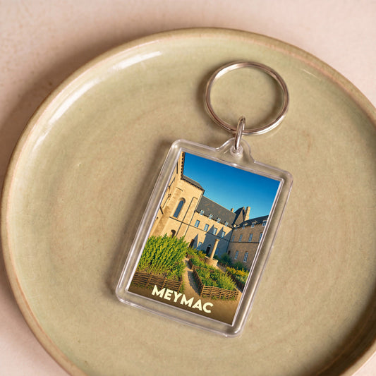 Porte-clés souvenir de la ville de Meymac avec une illustration stylisée de son abbaye et de ses jardins.
