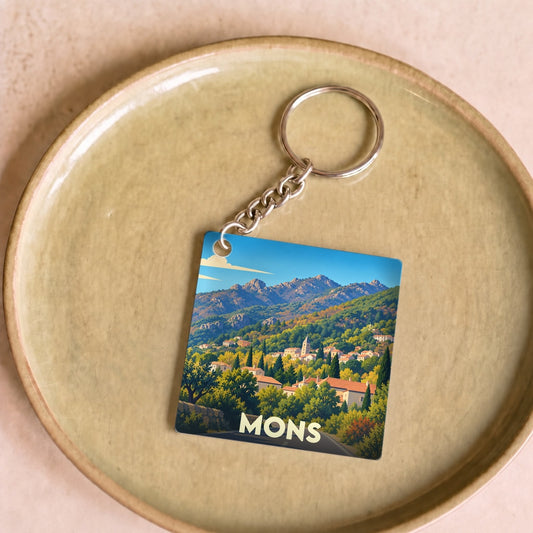 Porte-clés souvenir de la ville de Mons avec une illustration stylisée d'un village de montagne.