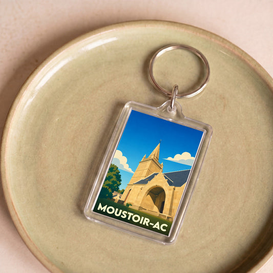 Porte-clés souvenir de la ville de Moustoir-Ac avec une illustration stylisée d'une église sous un ciel bleu.