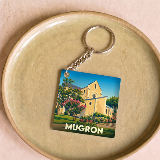 Porte-clés souvenir de la ville de Mugron avec une illustration stylisée de son église sous un ciel bleu.
