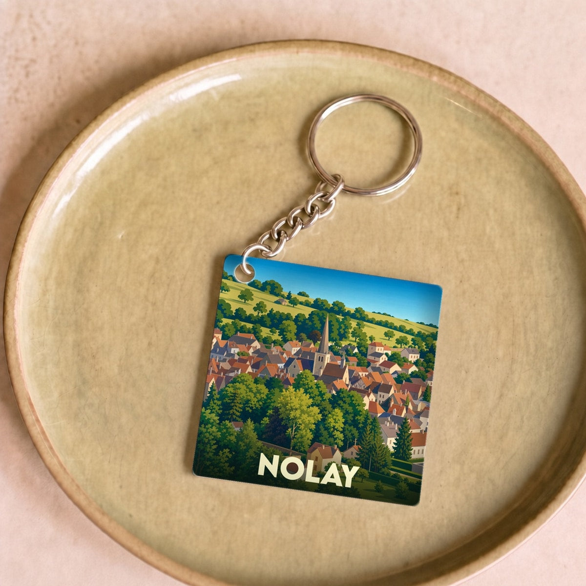 Porte-clés souvenir de la ville de Nolay avec une illustration stylisée d'un village dans une vallée verdoyante.