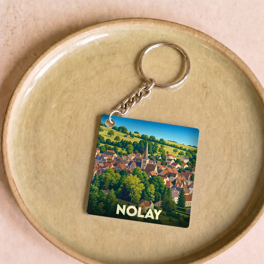 Porte-clés souvenir de la ville de Nolay avec une illustration stylisée d'un village dans une vallée verdoyante.