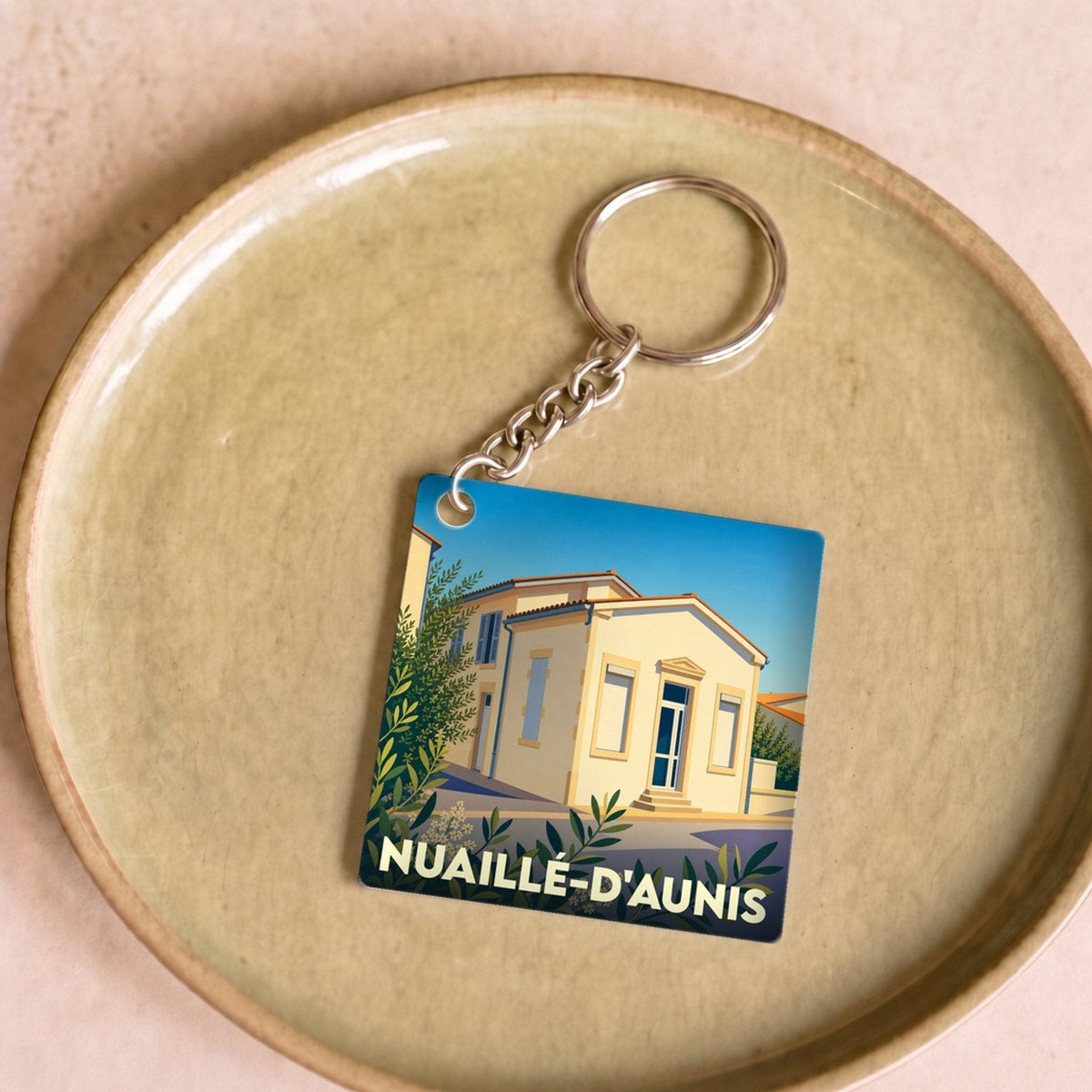Porte-clés souvenir de la ville de Nuaillé-d'Aunis avec une illustration stylisée d'un bâtiment sous un ciel bleu.