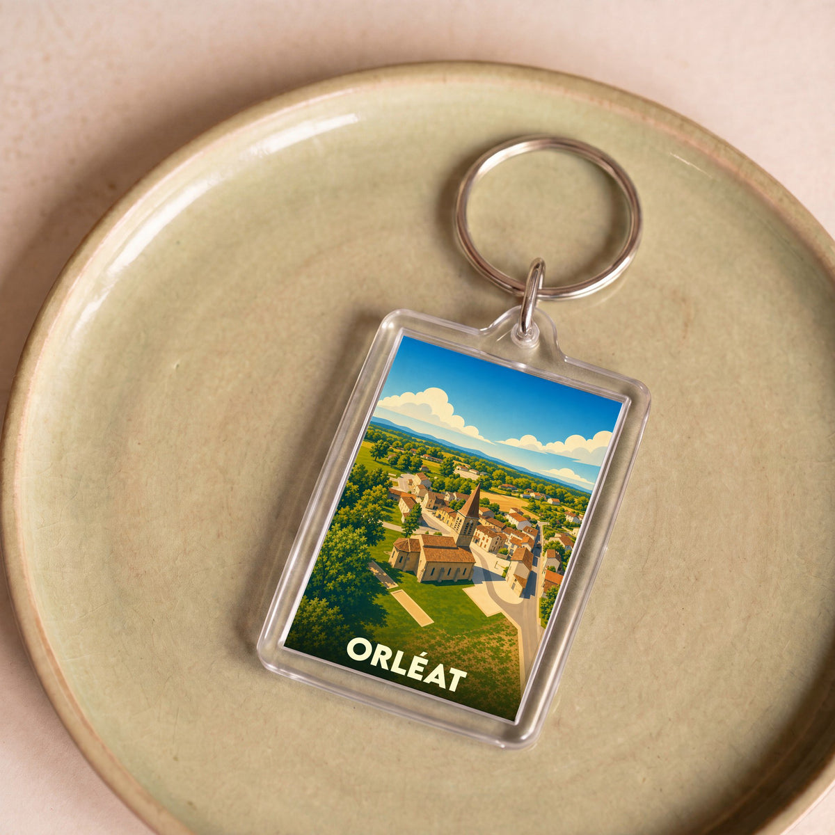 Porte-clés souvenir de la ville d'Orléat avec une illustration stylisée d'un paysage de village.