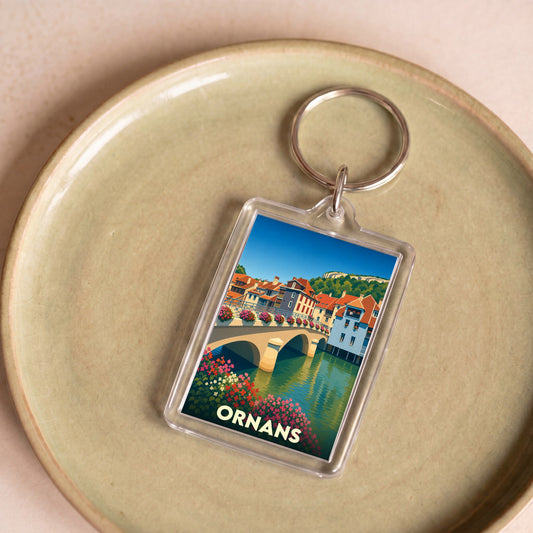 Porte-clés souvenir de la ville d'Ornans avec une illustration stylisée de son pont fleuri et de ses maisons au bord de l'eau.