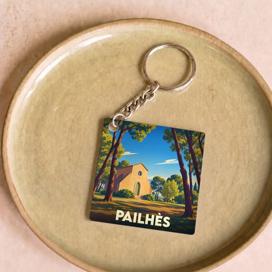Porte-clés souvenir de la ville de Pailhès avec une illustration stylisée d'une chapelle entourée de pins.