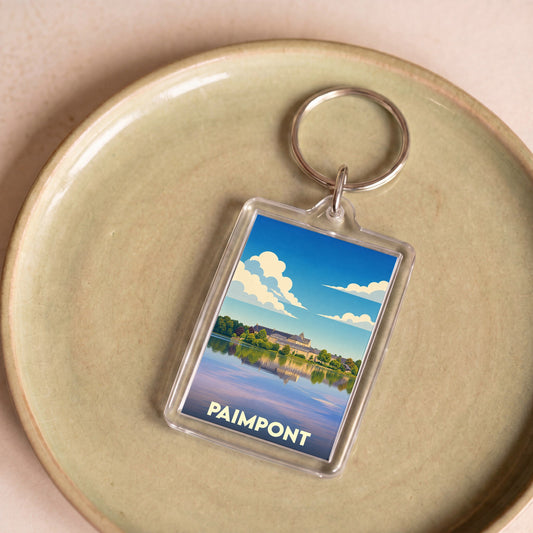 Porte-clés souvenir de la ville de Paimpont avec une illustration stylisée de son abbaye au bord de l'eau.