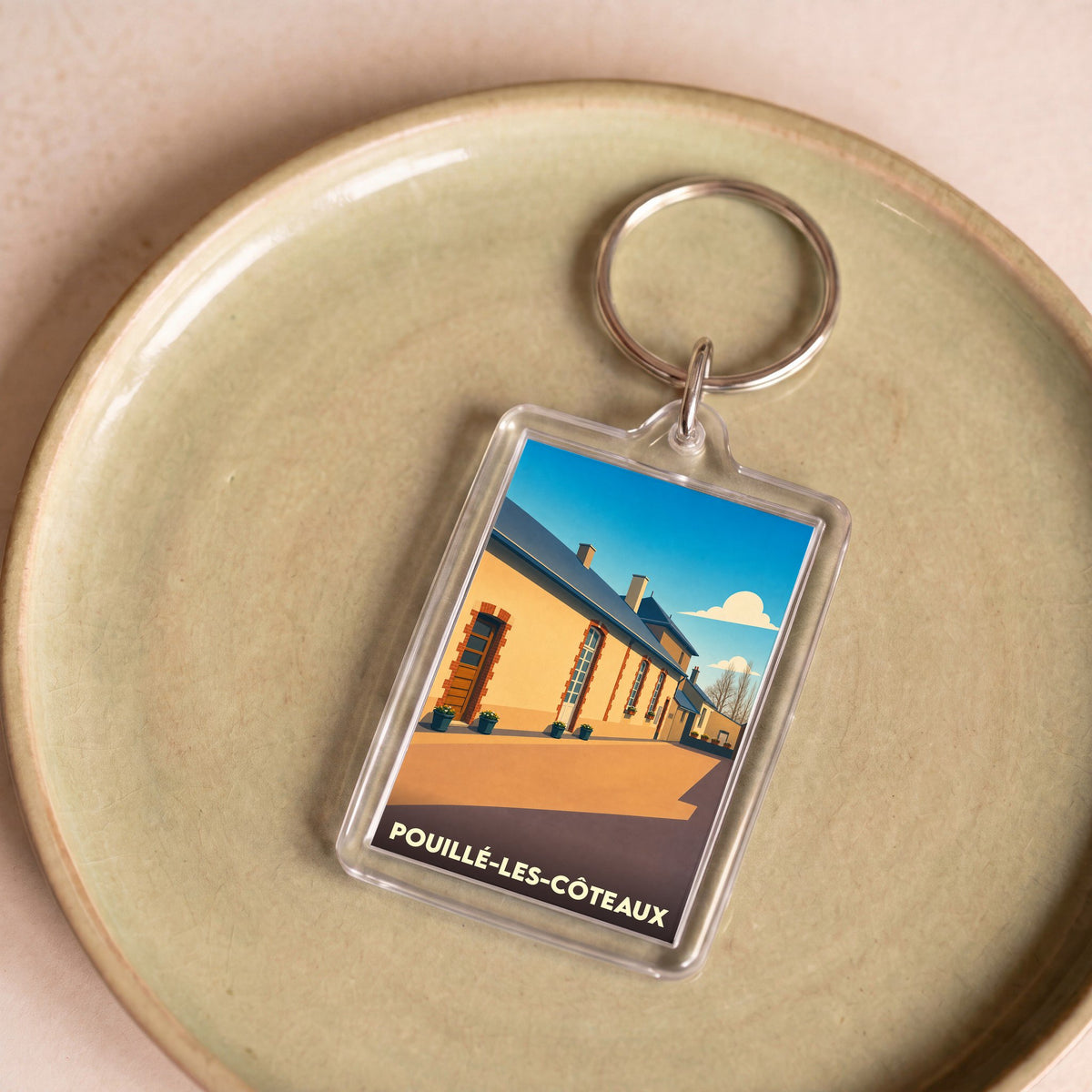 Porte-clés souvenir de la ville de Pouillé-les-Côteaux avec une illustration stylisée d'un bâtiment sous un ciel bleu.