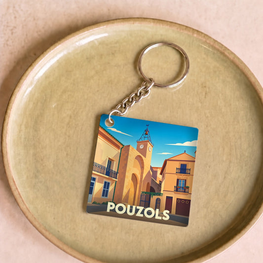 Porte-clés souvenir de la ville de Pouzols avec une illustration stylisée d'une rue typique et de son horloge.