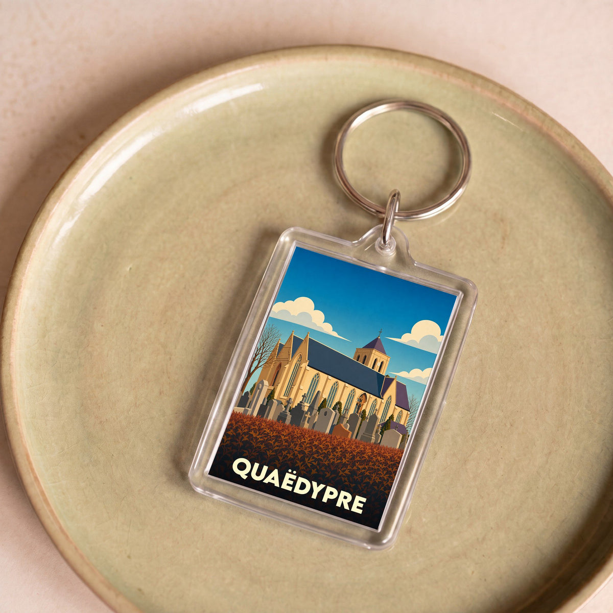 Porte-clés souvenir de la ville de Quaëdypre avec une illustration stylisée de son église et de son cimetière.