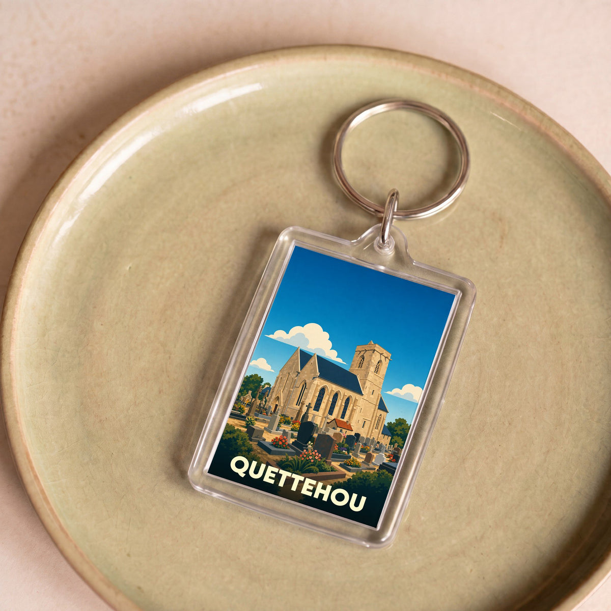 Porte-clés souvenir de la ville de Quettehou avec une illustration stylisée de son église et de son cimetière.