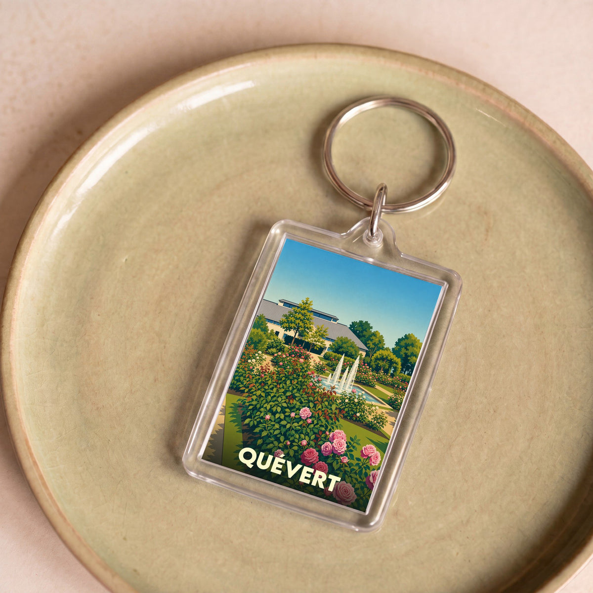 Porte-clés souvenir de la ville de Quévert avec une illustration stylisée d'un jardin fleuri et de ses fontaines.