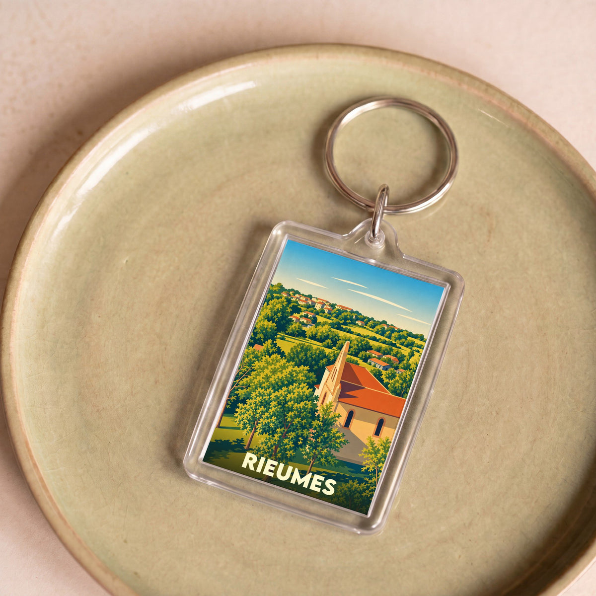 Porte-clés souvenir de la ville de Rieumes avec une illustration stylisée d'un paysage de campagne et de son église.