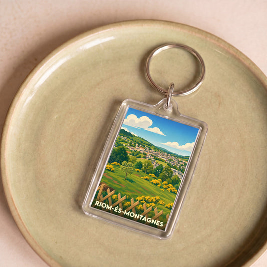 Porte-clés souvenir de la ville de Riom-ès-Montagnes avec une illustration paysagère stylisée.