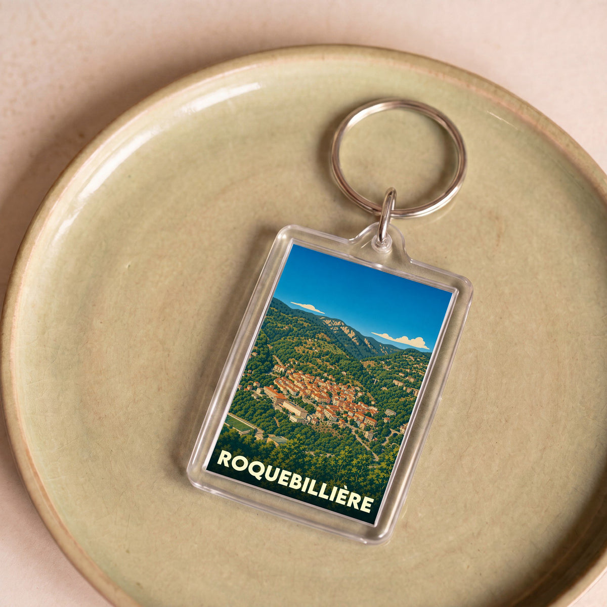 Porte-clés souvenir de la ville de Roquebillière avec une illustration stylisée du village dans les montagnes.