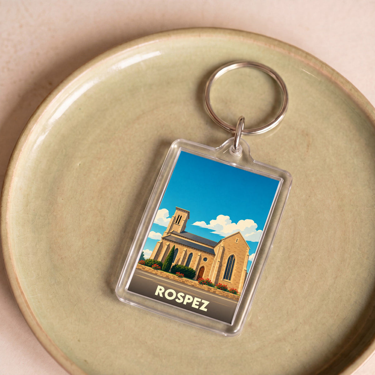 Porte-clés souvenir de la ville de Rospez avec une illustration stylisée de son église sous un ciel bleu.