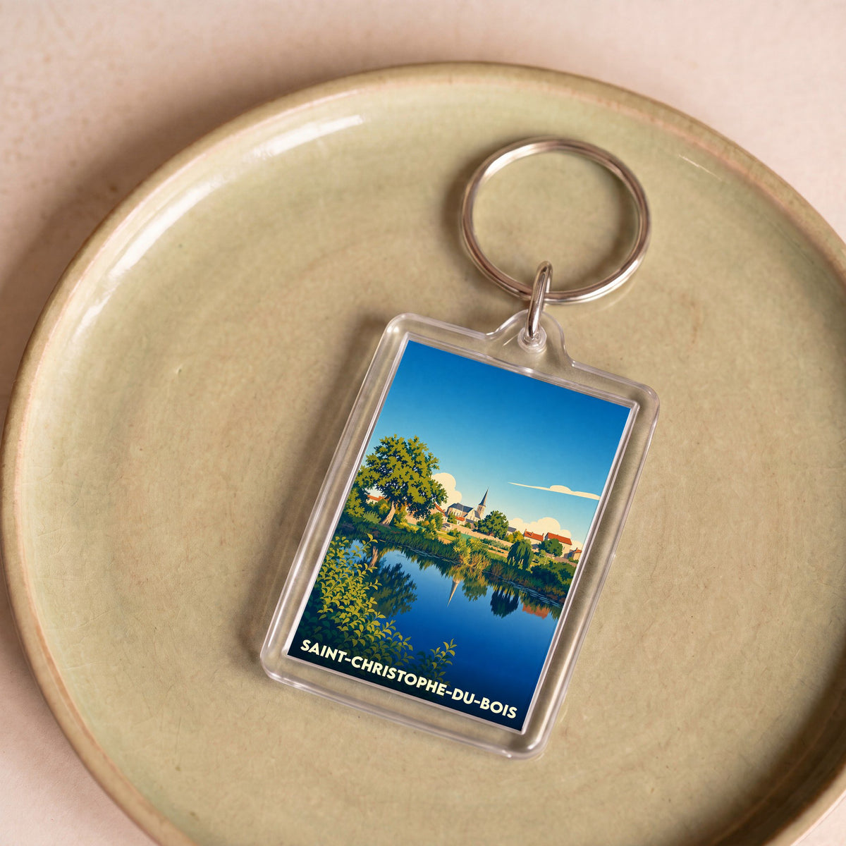 Porte-clés souvenir de la ville de Saint-Christophe-du-Bois avec une illustration stylisée d'un paysage au bord de l'eau.