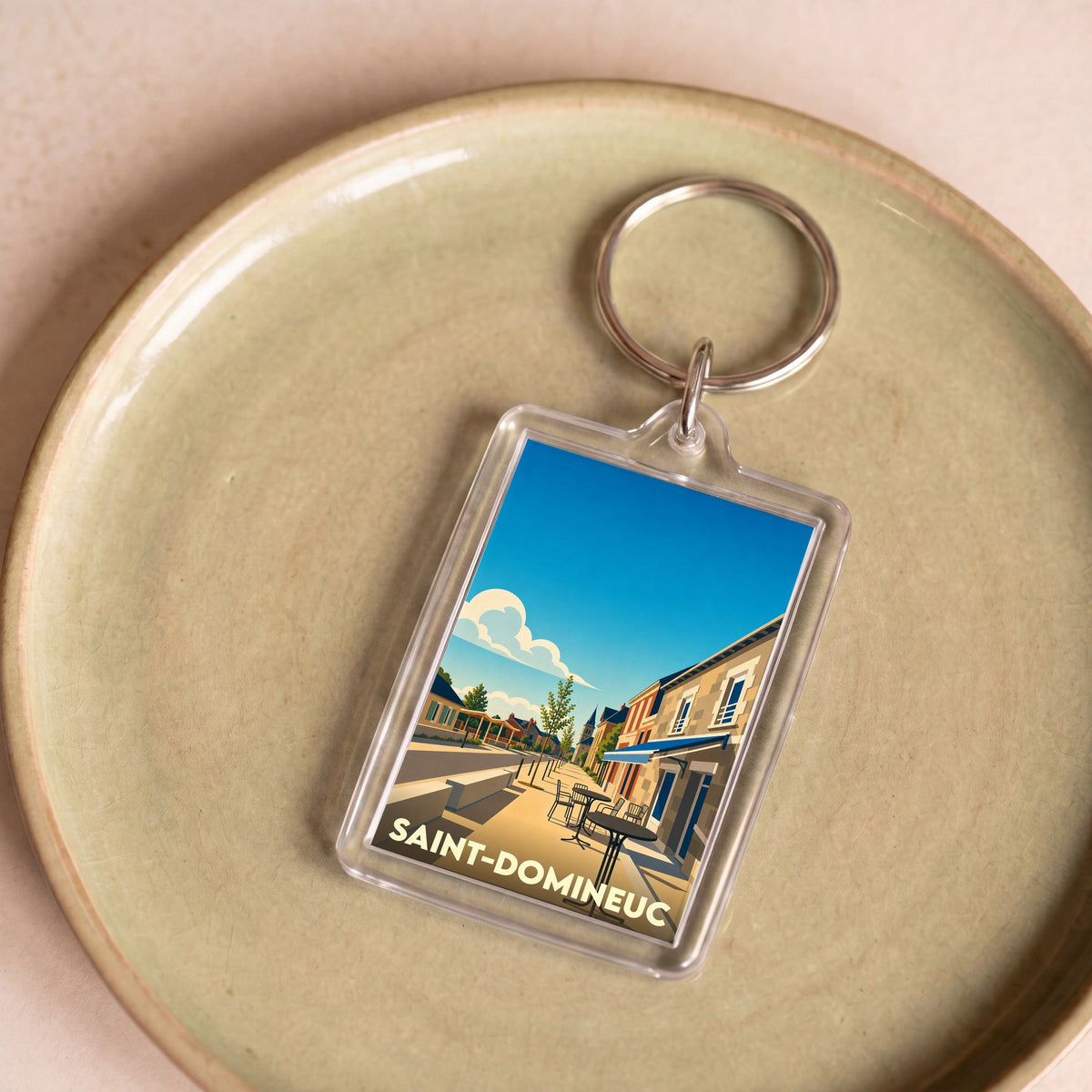Porte-clés souvenir de la ville de Saint-Domineuc avec une illustration stylisée d'une rue ensoleillée.