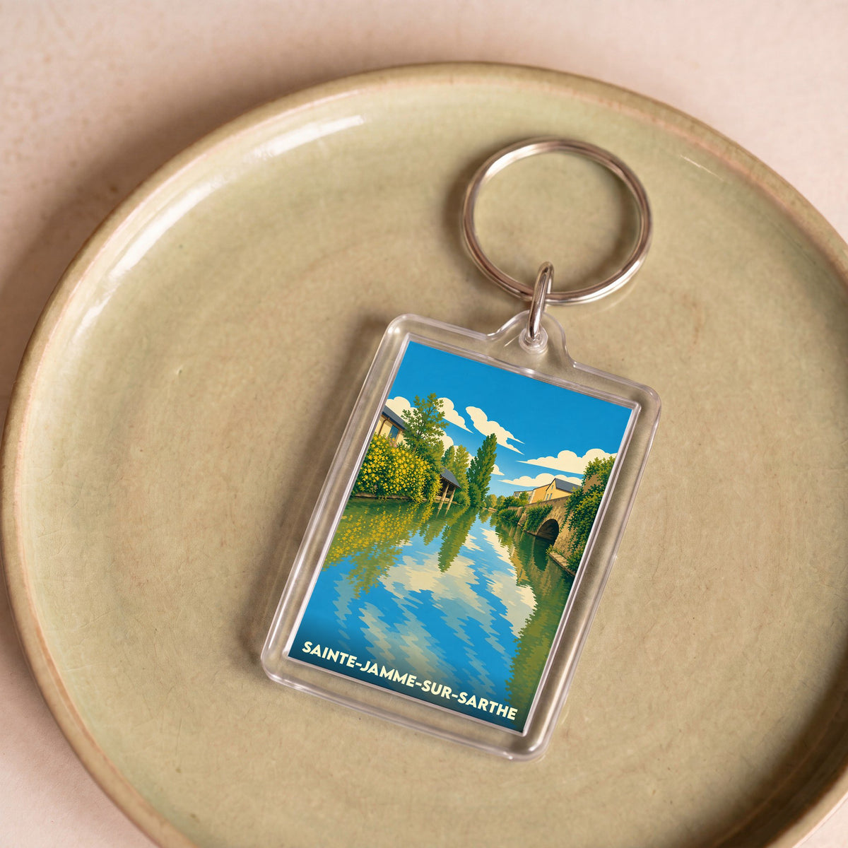 Porte-clés souvenir de la ville de Sainte-Jamme-sur-Sarthe avec une illustration stylisée d'un cours d'eau bordé de verdure.