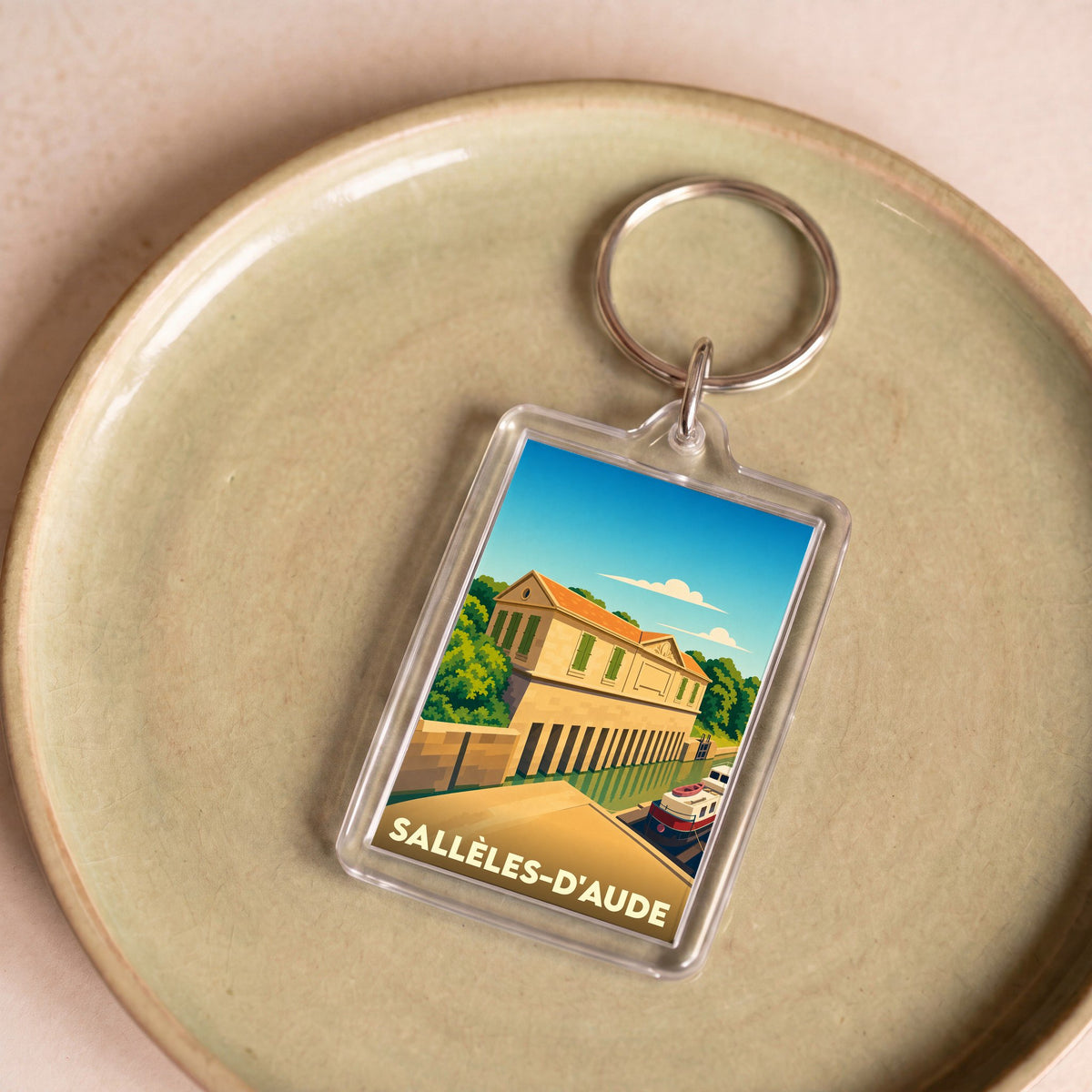 Porte-clés souvenir de la ville de Sallèles-d'Aude avec une illustration stylisée d'un bâtiment au bord de l'eau.
