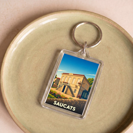 Porte-clés souvenir de la ville de Saucats avec une illustration stylisée d'une maison bourgeoise locale.