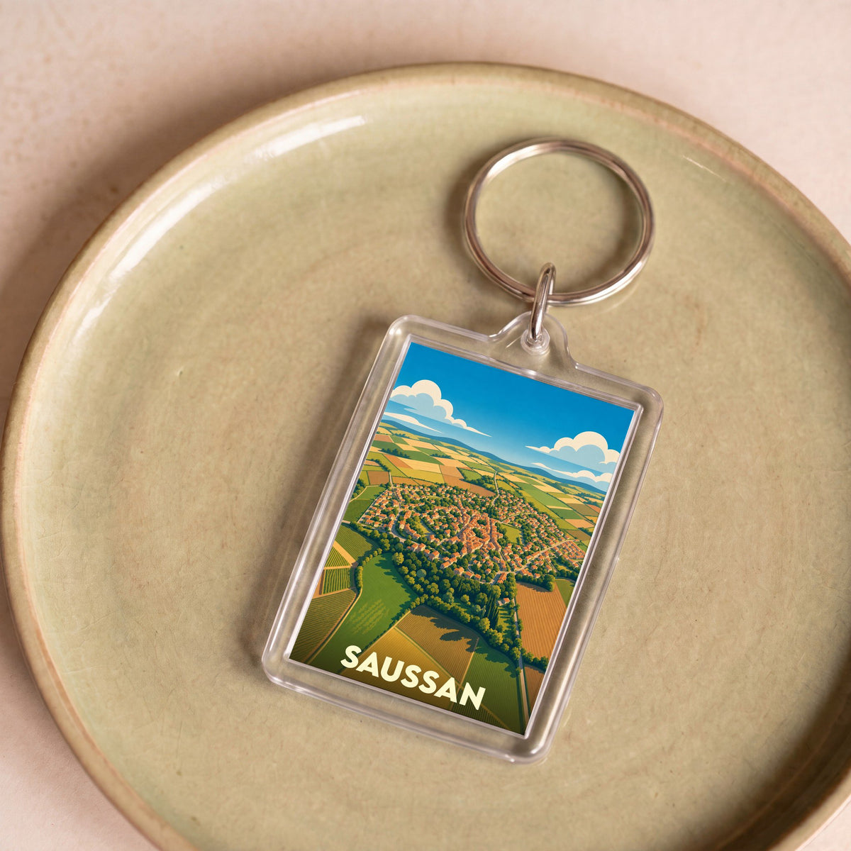 Porte-clés souvenir de la ville de Saussan avec une illustration aérienne stylisée du village et de ses champs.