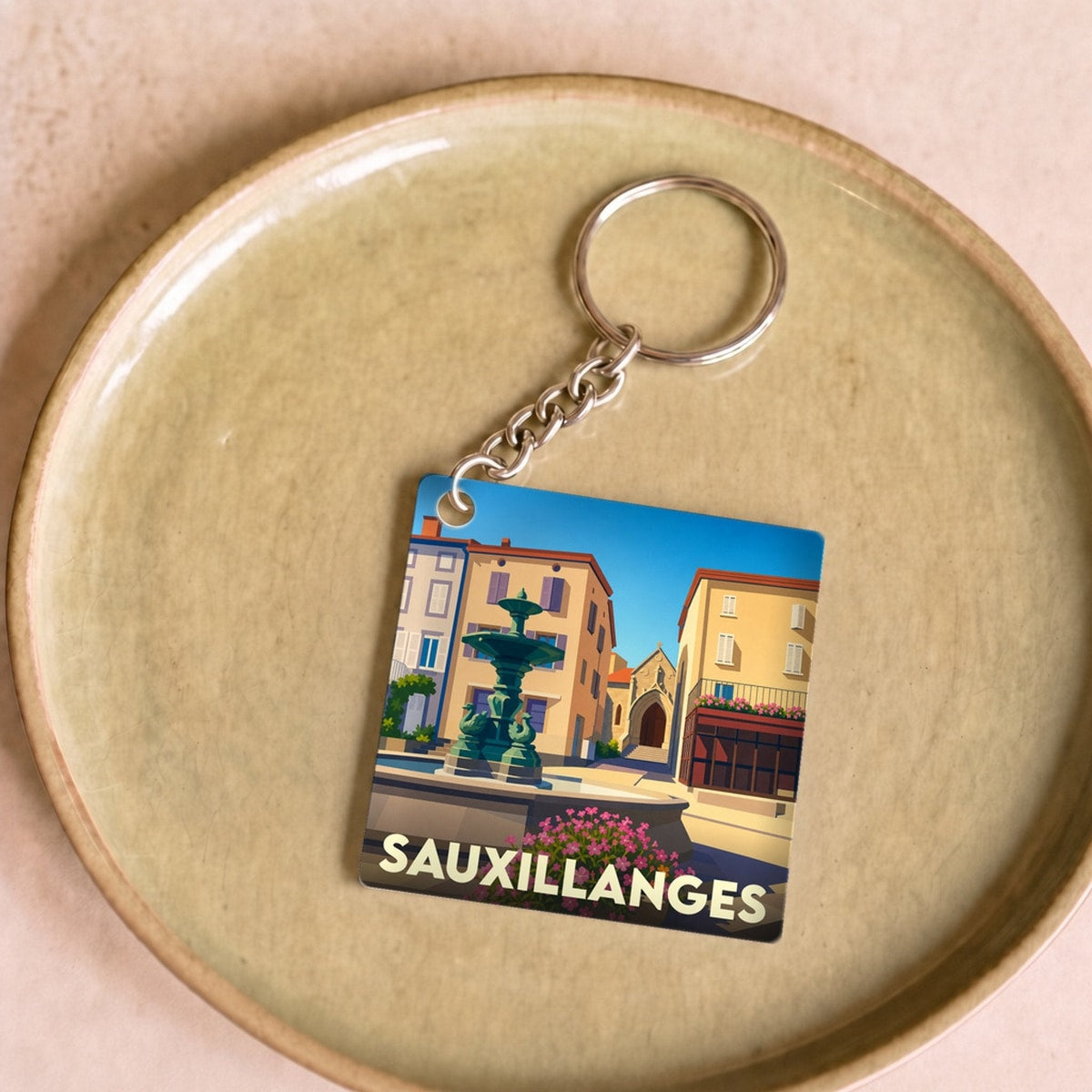 Porte-clés souvenir de la ville de Sauxillanges avec une illustration stylisée d'une place et sa fontaine.