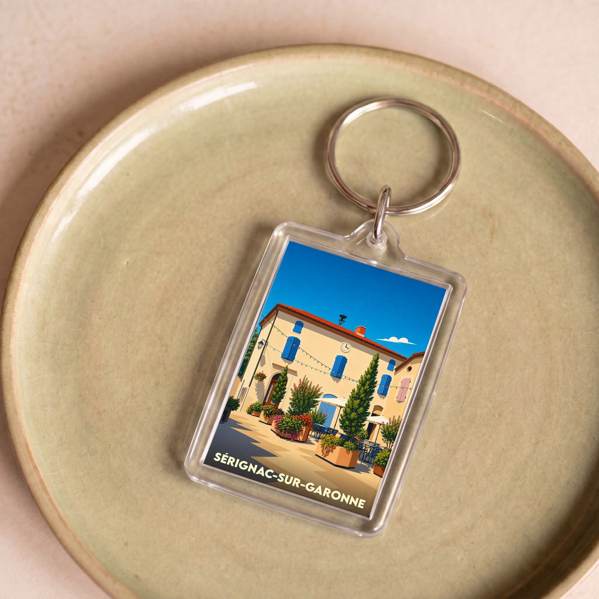 Porte-clés souvenir de Sérignac-sur-Garonne avec une illustration stylisée d'une place ensoleillée.