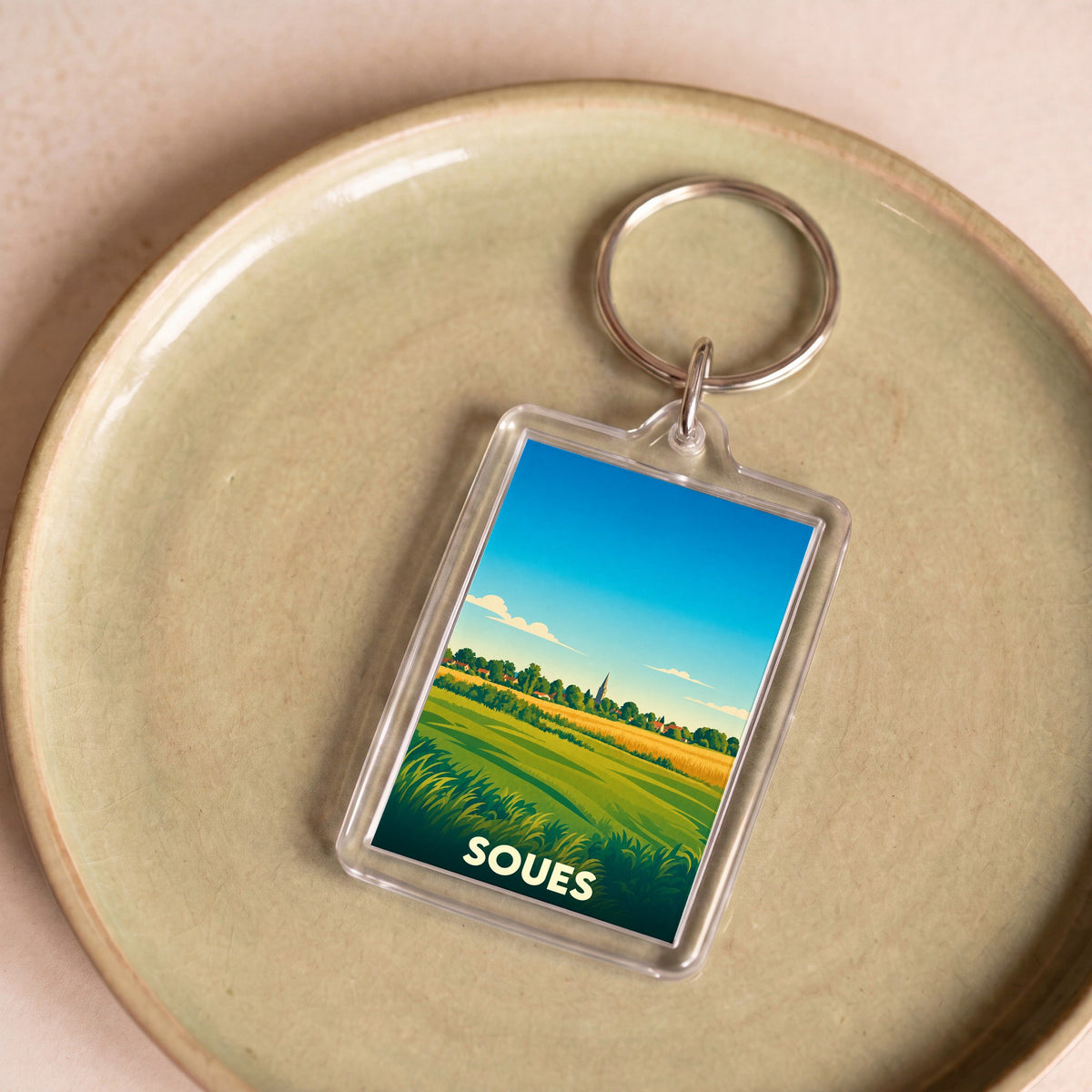 Porte-clés souvenir de la ville de Soues avec une illustration stylisée de paysages champêtres.