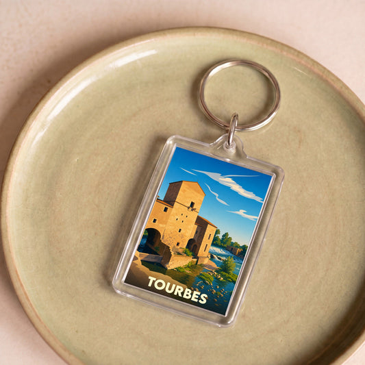 Porte-clés souvenir de la ville de Tourbes avec une illustration stylisée d'un bâtiment historique au bord de l'eau.