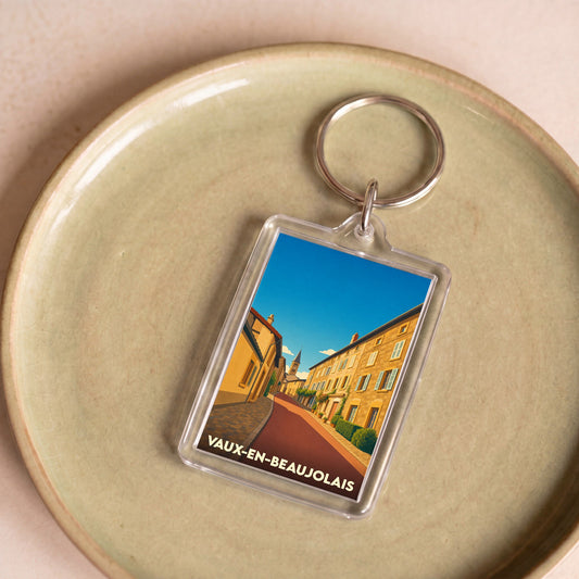 Porte-clés souvenir de la ville de Vaux-en-Beaujolais avec une illustration stylisée d'une rue ensoleillée.